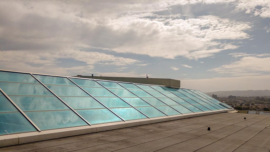 Rooftop with a skyligt with angled, large blue-tinted glass panels under a partly cloudy sky, overlooking a distant cityscape. Calm and serene atmosphere.
