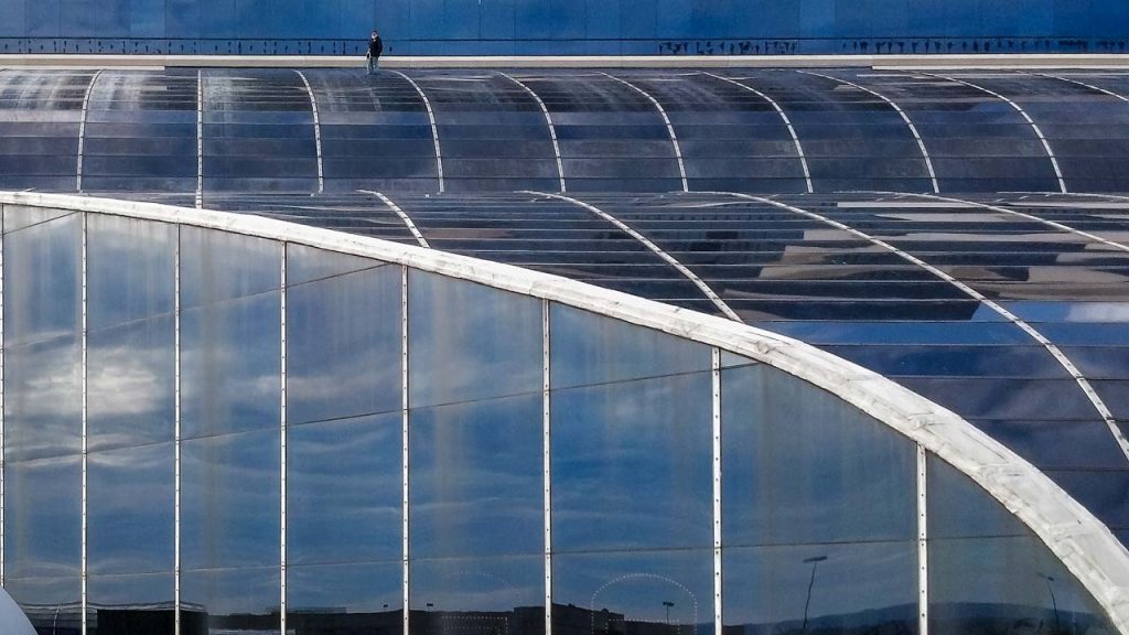 A lone person walks across a large, curved glass roof. The reflective panels create a geometric pattern, mirroring a cloudy sky, conveying isolation.
