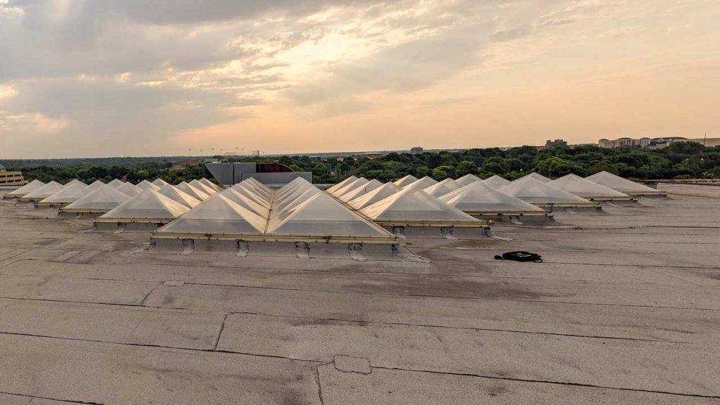 Skylight pyramid domes arranged in rows on a large, flat rooftop under a cloudy sky at sunset, with trees and distant buildings on the horizon.