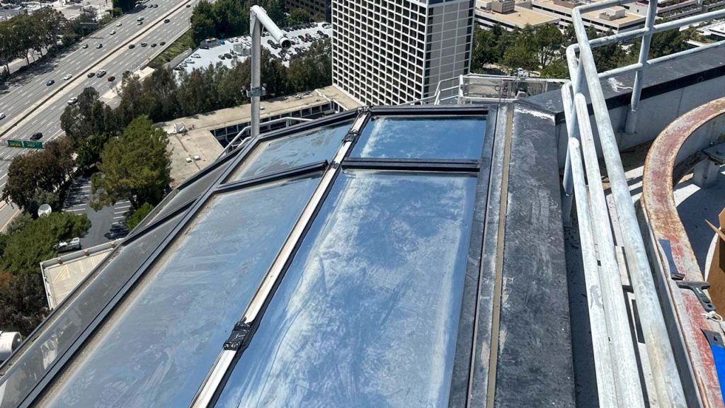 View from a rooftop showing reflective skylights, safety railings, and nearby buildings under a clear blue sky. A highway runs below, bordered by trees.
