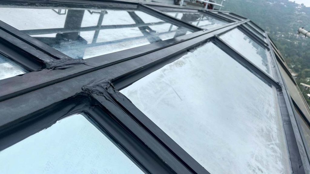 A close-up view of dark sealant or caulking freshly applied along the black metal joints and framing of a commercial glass skylight.