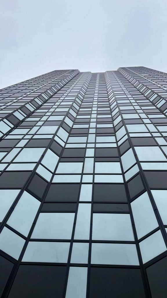 A tall skyscraper viewed from below, with a grid of reflective glass windows stretching upward into a gray, overcast sky, conveying a sense of modernity.