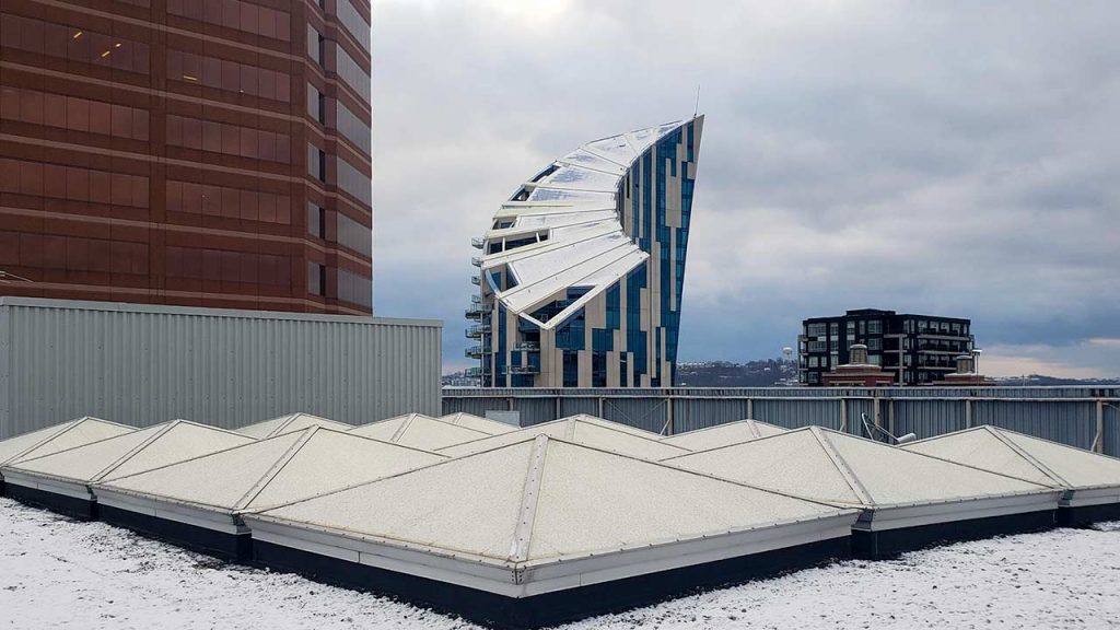 Snow-covered rooftop with a cluster of , pyramid skylights. A gray, overcast sky and urban buildings in the background convey a cold, wintry atmosphere.