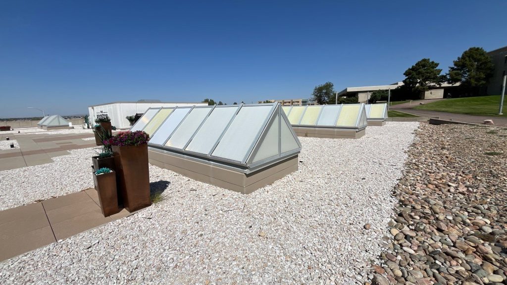 An image showing three ridge skylights is installed on a gravel rooftop. The background shows grass, trees, and a beige building.