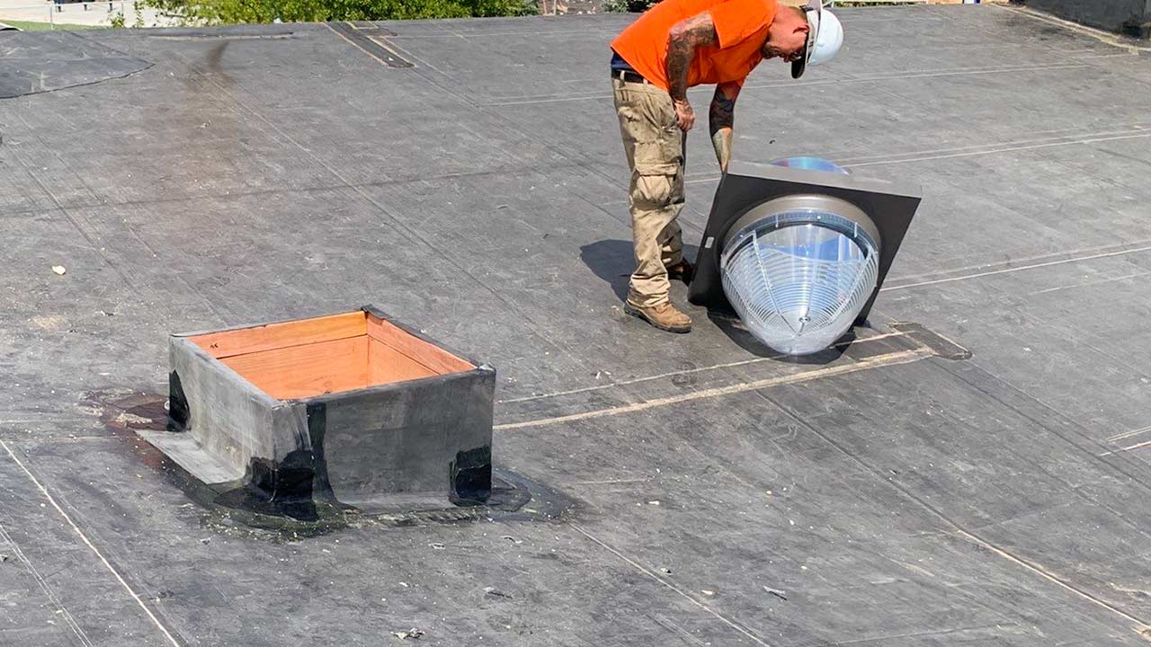 A worker in an orange shirt and hard hat installs a Sun Tunnel or light tube on a flat, black rubber roof next to an unfinished roof opening.