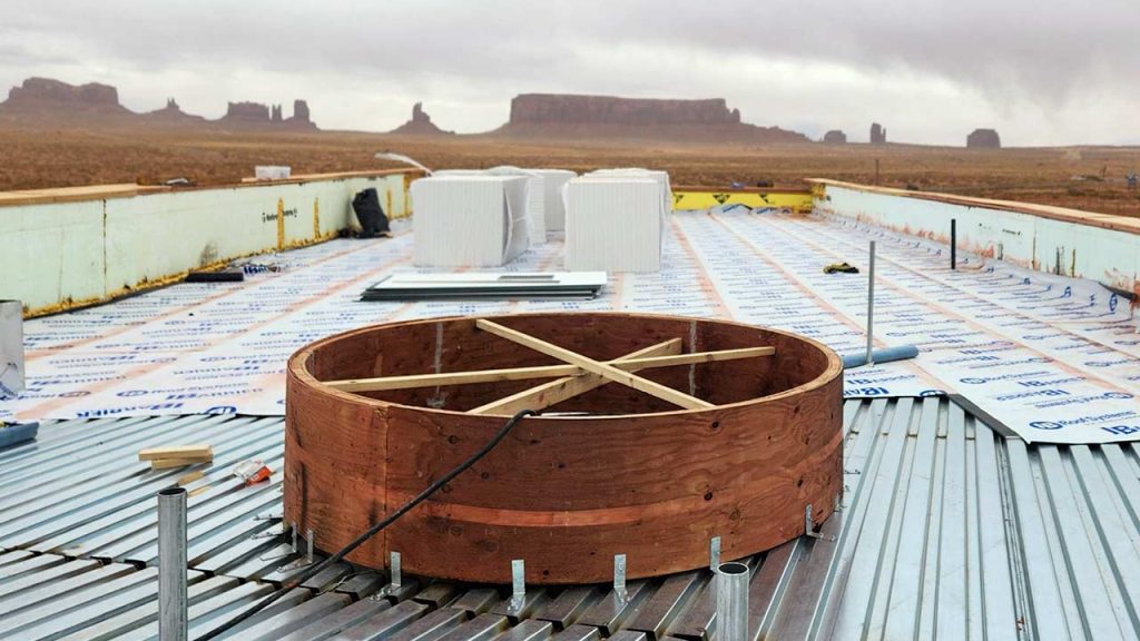 A rooftop under construction with insulation sheets, HVAC units, and a large wooden circular skylight curb , set against a desert landscape with distant mesas.
A rooftop under construction with insulation sheets, HVAC units, and a large wooden circular frame, set against a desert landscape with distant mesas.