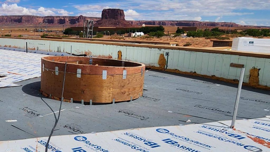A rooftop under construction with insulation sheets, and a large wooden circular skylight curb , set against a desert landscape with distant mesas.