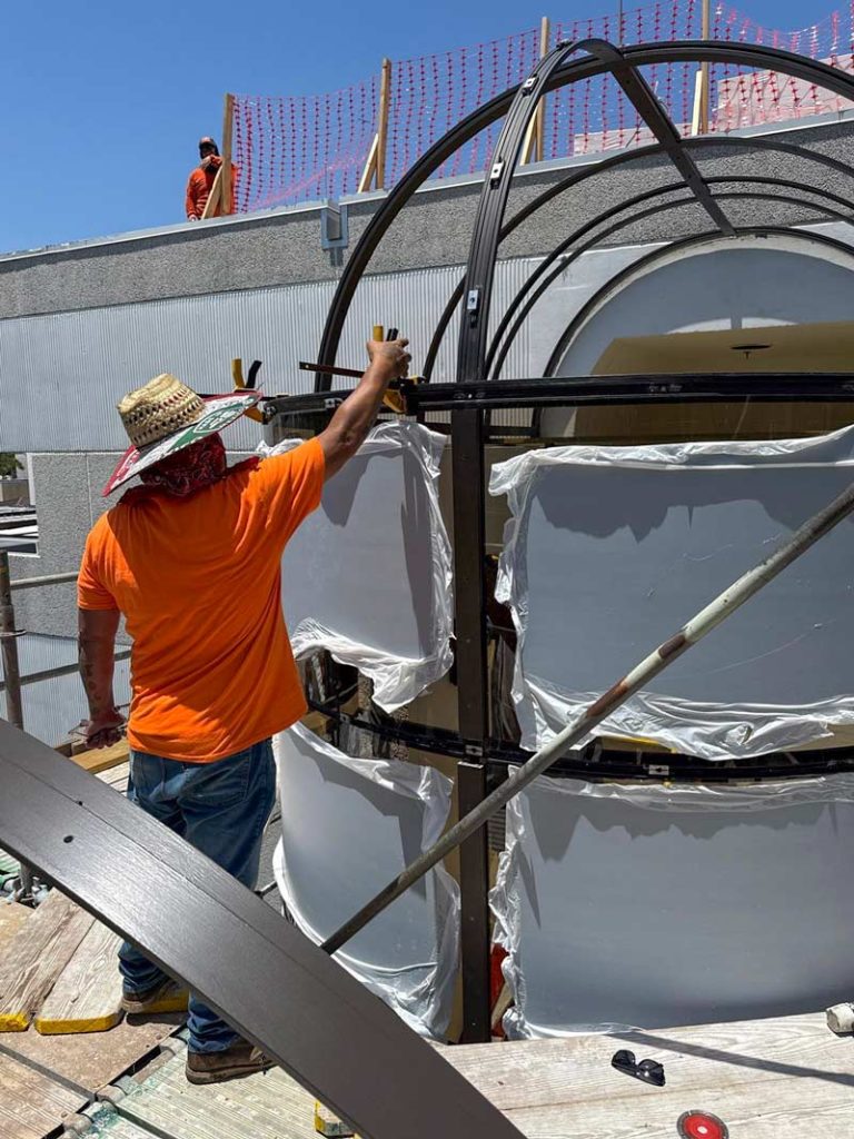 A man wearing an orange shirt works on replacing skylight panels in a large metal stairwell structure.