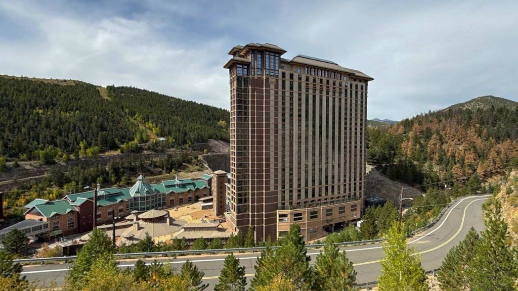 Tall 34-story brown hotel building, home to the Ameristar Casino, stands surrounded by trees and mountains, next to a winding road under a cloudy sky.