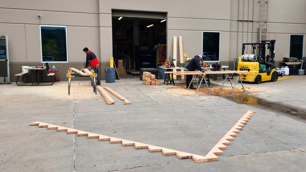 Outdoor workspace with two people working on wooden stair risers on sawhorses. A forklift and wood pieces are nearby. The scene conveys industrious activity.