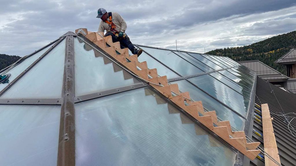 A worker in safety gear works on wooden stair risers on a large glass skylight, set against a backdrop of a cloudy sky and forested hills.