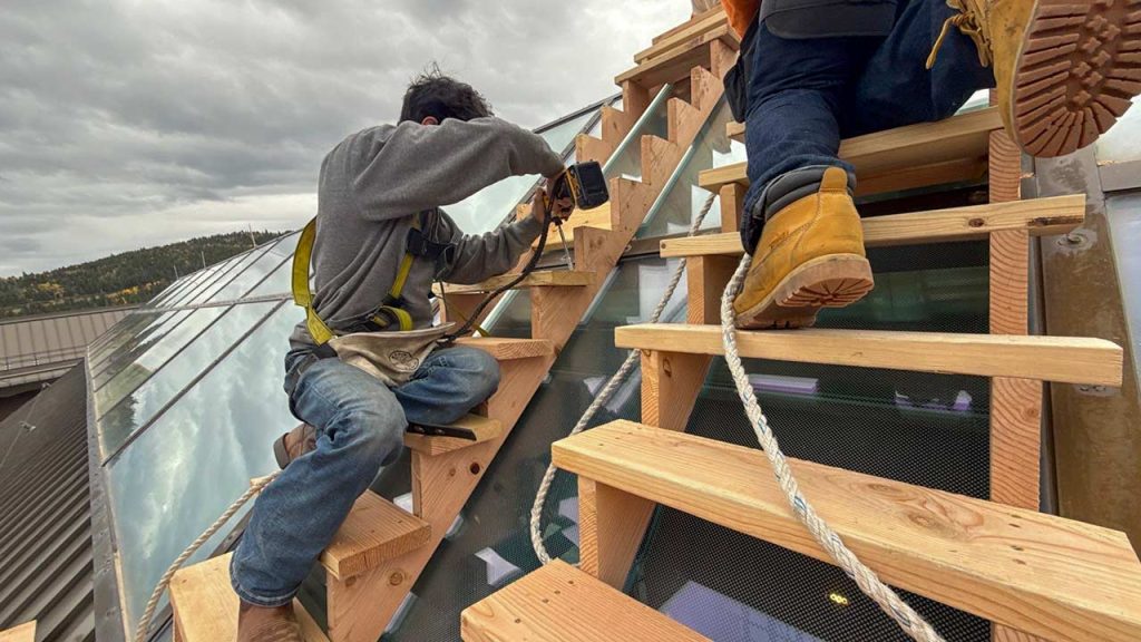 Two construction workers in safety gear are attaching wooden stairs above a glass skylight, using tools under a cloudy sky. The scene conveys focus and teamwork.