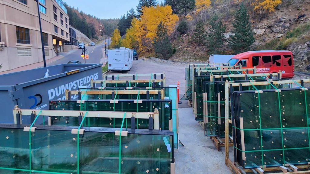 Stacks of glass units are secured with straps and wooden frames in a parking lot. A large blue trash container and a red van are nearby. Autumn trees line the background.
