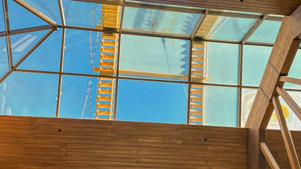 View through a glass skylight shows a clear blue sky and a wooden outdoor staircase. Wooden beams frame the scene, creating a modern, airy atmosphere.