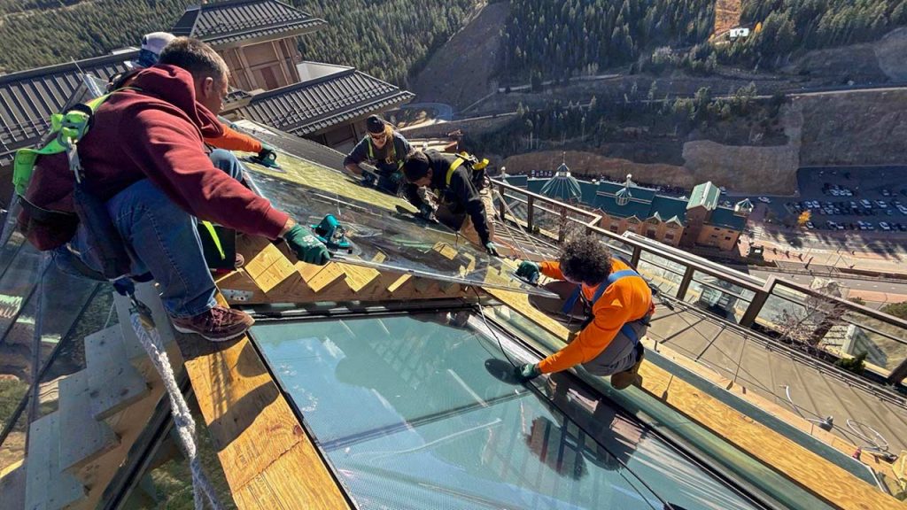 Workers in safety gear install glass units on a steep rooftop skylight with mountain views. The scene conveys teamwork, precision, and a sense of height.