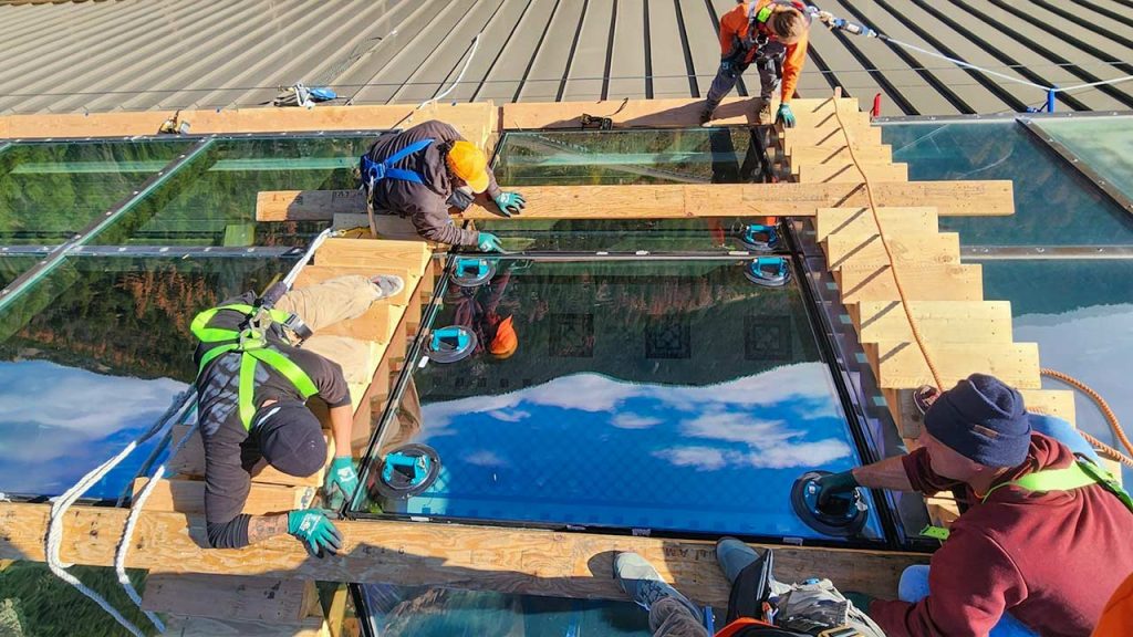 Workers wearing safety gear are installing a large glass unit on a skylight. The glass reflects blue skies and clouds, conveying a sense of openness.