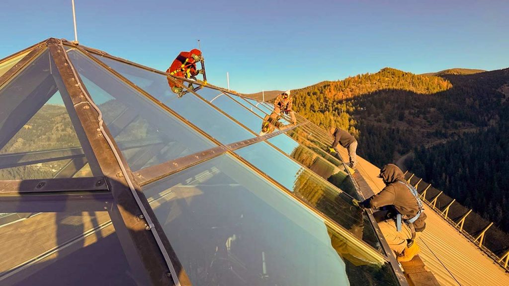 Workers in safety gear clean a large glass extended yramid skylight, reflecting a scenic mountain backdrop. The mood is diligent and serene.