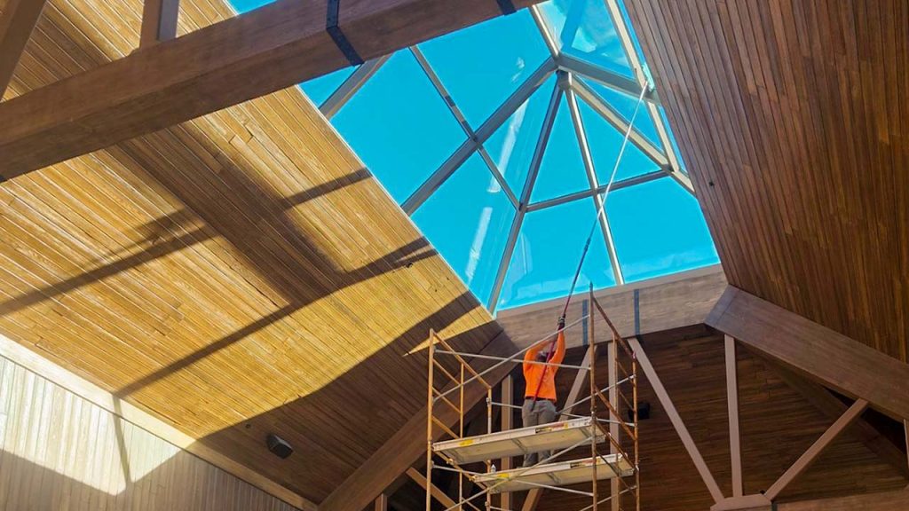 A worker in an orange vest stands on scaffolding, cleaning a large, extended pyramid glass skylight. Sunlight creates dramatic shadows on wooden walls.