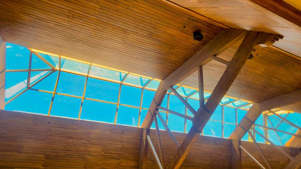 Wooden ceiling with large triangular beams and a large atrium skylight. Sunlight streams through, creating a warm, airy, and spacious atmosphere.