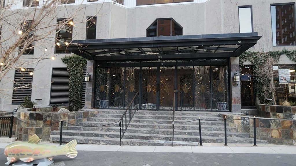 Entrance of a modern building with metal and glass doors, surrounded by stone walls and greenery. A large fish sculpture is in the foreground, adding an artistic touch.