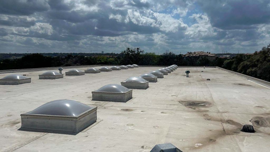 Flat rooftop with rows of glossy, dome-shaped skylights under a partly cloudy sky. The landscape in the distance is dotted with trees.