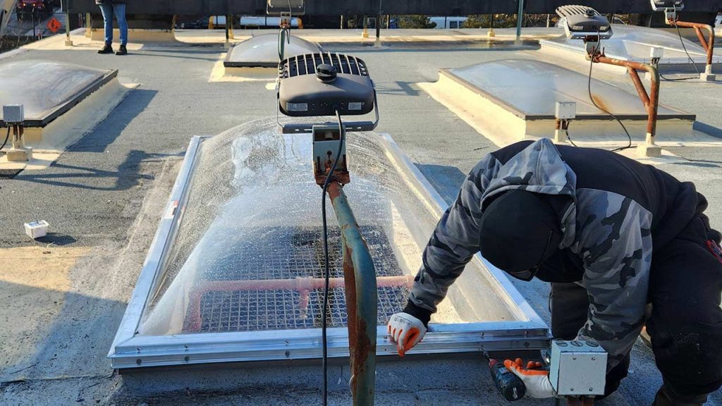 A worker in a camouflage jacket installs a skylight on a rooftop. Nearby are skylights and lighting units. The scene conveys focus and precision.