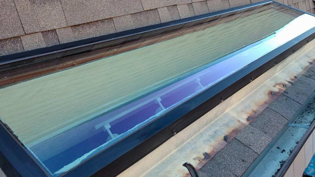A large skylight on a shingled roof reflects colorful hues of blue, green, and purple. The surrounding roof appears weathered, showing signs of rust.