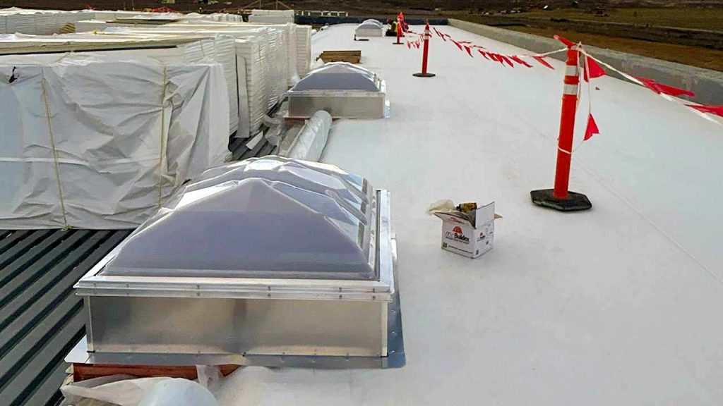 A flat white rooftop under construction features two large domed skylights and stacked building materials. Safety cones and red flags line the perimeter.