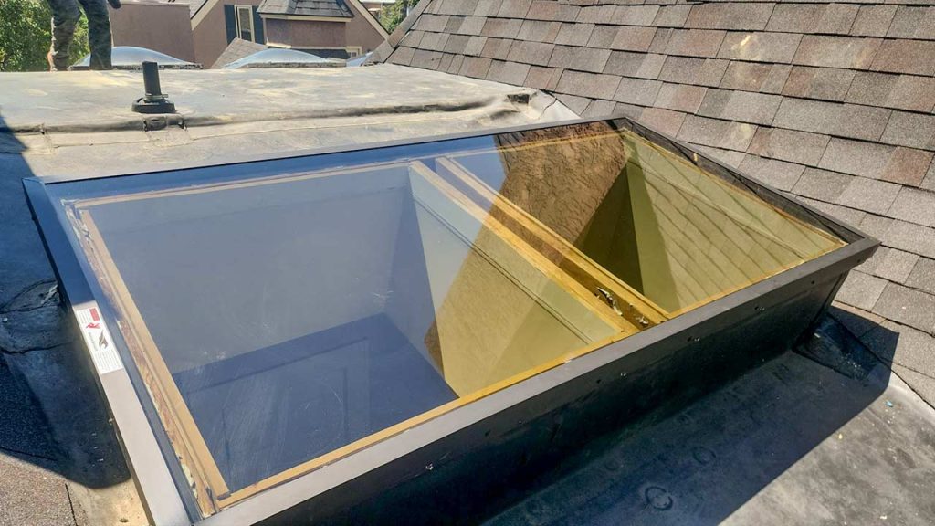 A rooftop skylight with a clear glass window set in a dark metal frame, surrounded by gray shingles under bright sunlight, creating a modern look.