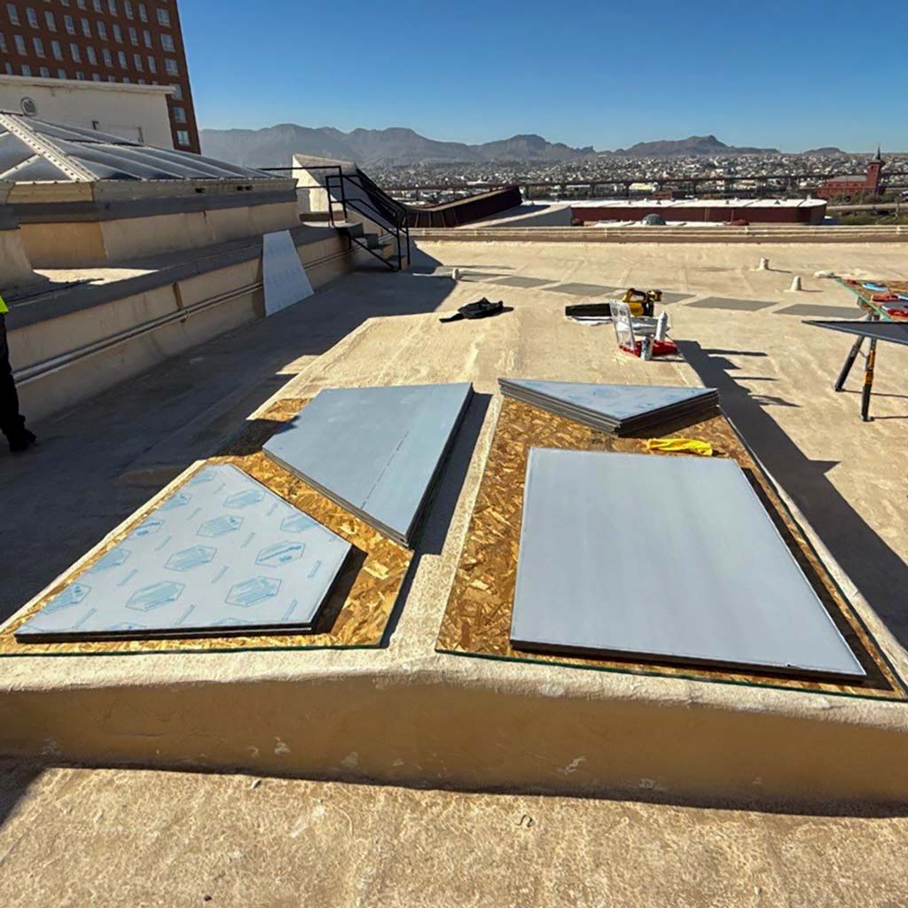 Rooftop view with mountain backdrop showing sheets of Polycarvonate skylight panels laid out. Clear blue sky, construction tools, and larger buildings nearby.