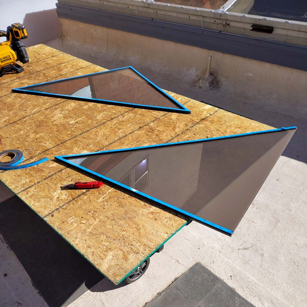 Two triangular polycarbonate panes with blue gaskets lie on a wooden table outdoors. A yellow drill and a red utility knife are nearby, suggesting ongoing construction.