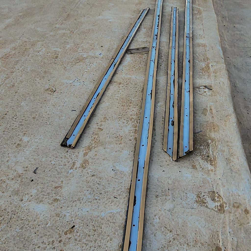 Four metal strips with holes lie on a textured roofing surface. The items are weathered and aligned parallel, conveying an industrial tone.