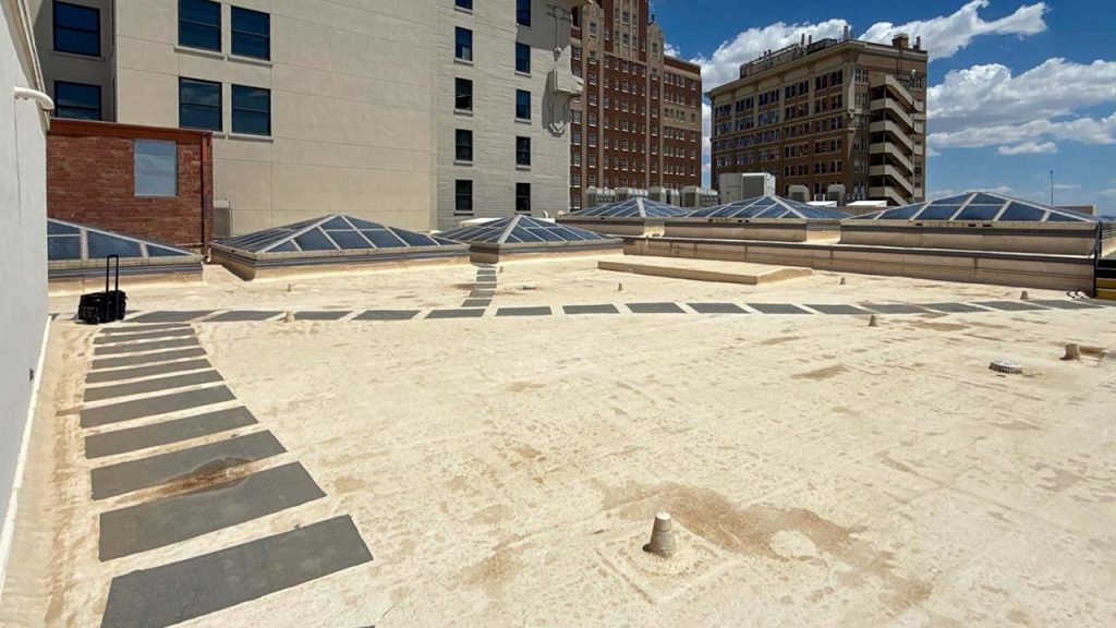 A sunny rooftop scene features beige tiles, with rows of pyramid skylights. Background shows tall, urban buildings under a blue sky with scattered clouds.