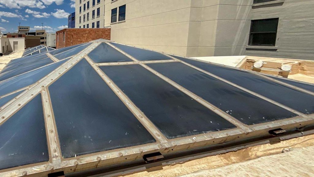 Pyramid skylight structure on a building roof with a clear blue sky and scattered clouds. Surrounding buildings suggest an urban setting.