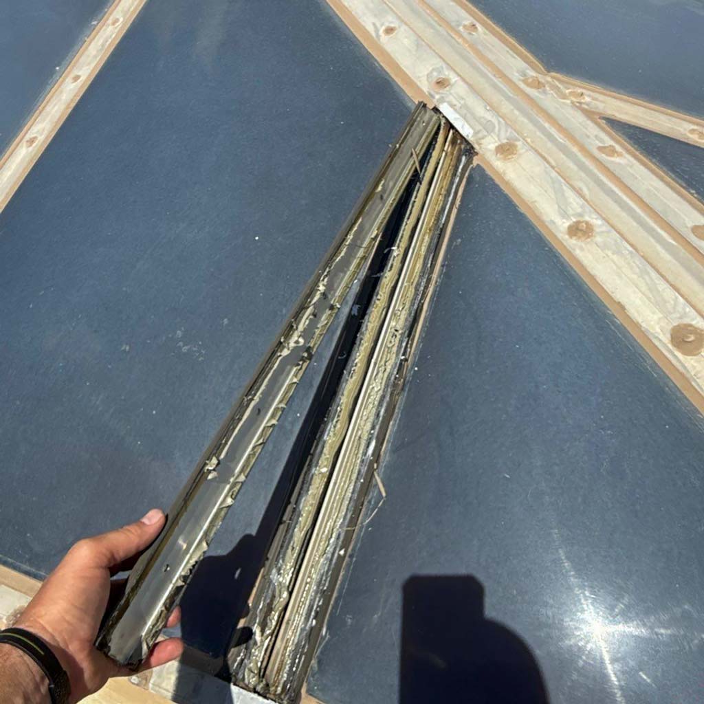 A hand holds a weathered,metal piece against a background of polymer skylight panels, suggesting repair work. Bright sunlight creates strong shadows.