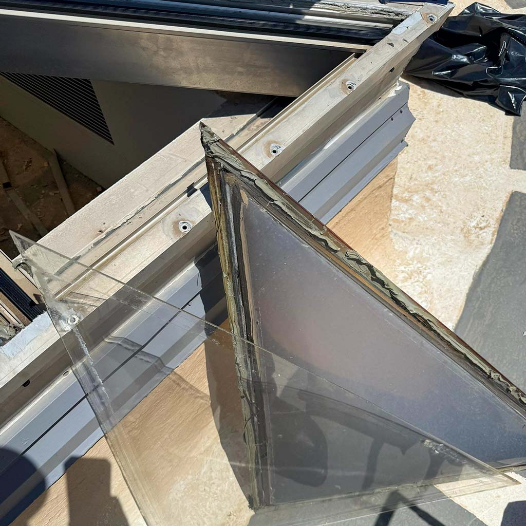 Close-up of a triangular plastic panel removed from a skylight frame, revealing an outdoor area. Sunlight casts shadows, evoking a sense of repair.