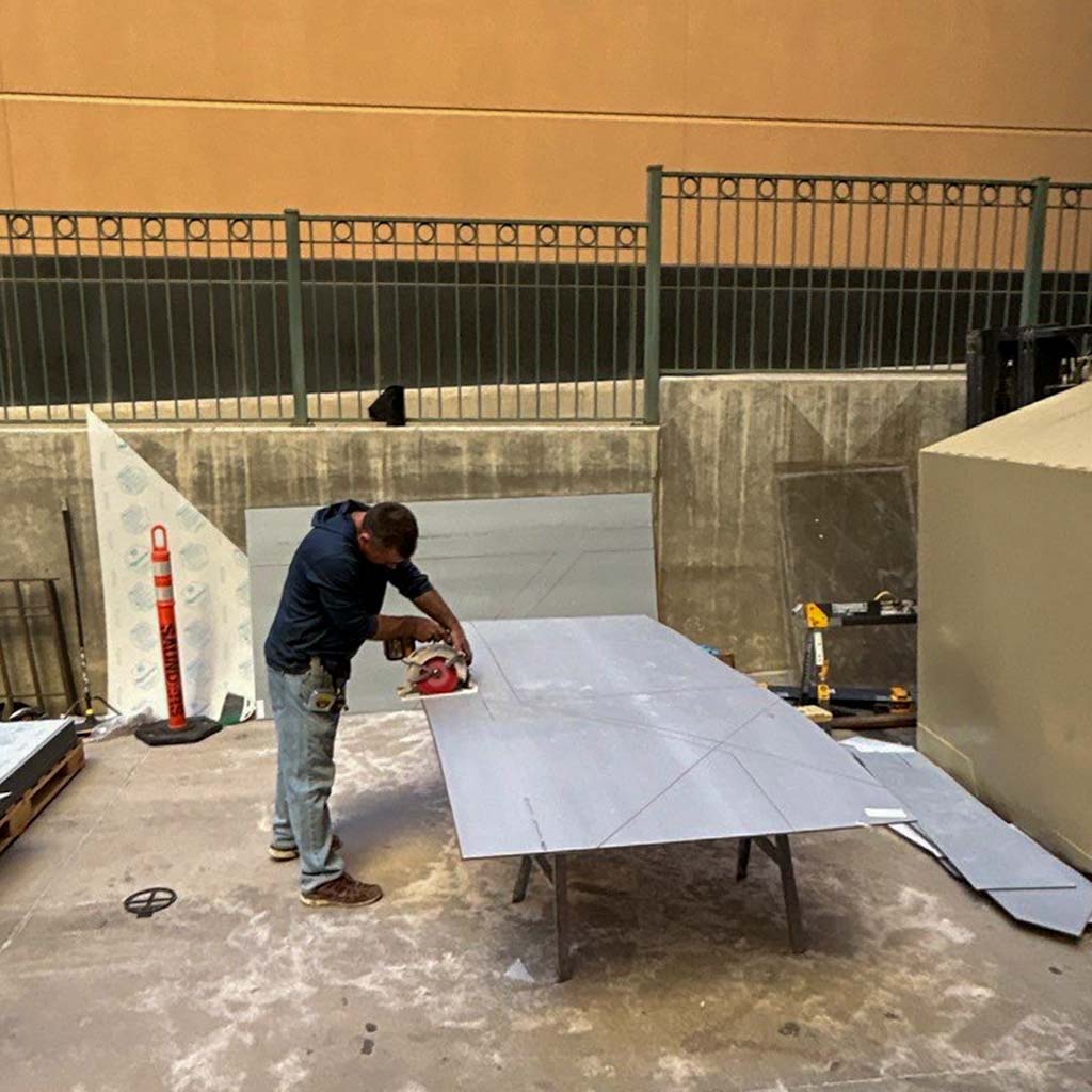 A man in blue clothing uses a circular saw to cut a large sheet of material on a table in an outdoor work area, surrounded by construction tools.