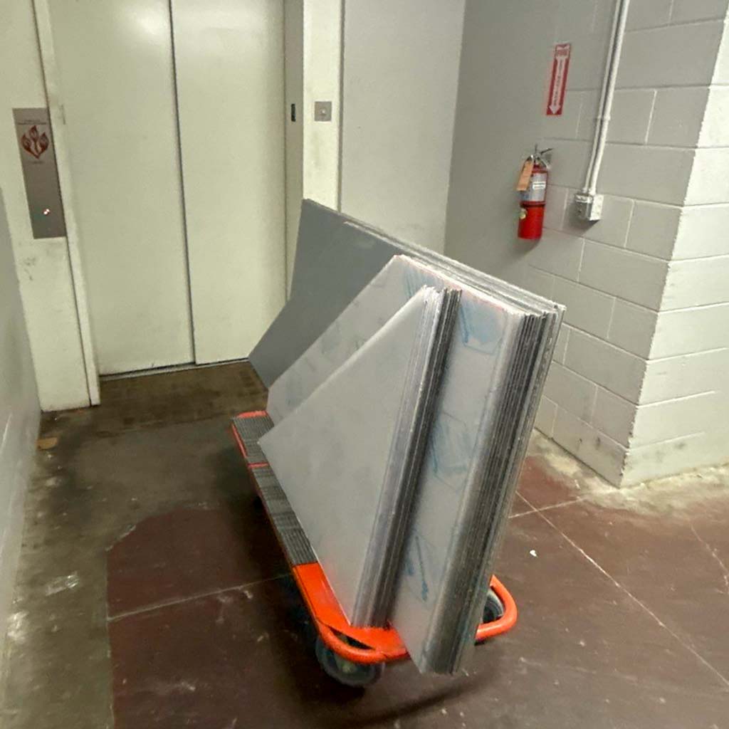 A cart loaded with large triangular and rectangular panels is positioned in a dimly lit building corridor, near an elevator and fire extinguisher.