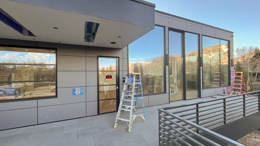 A modern building exterior with large glass windows and a door. Several ladders lean against the structure, indicating ongoing construction work.
