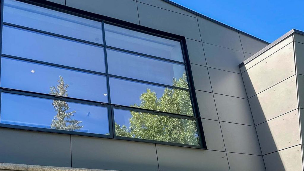 Modern gray building facade with large reflective windows showcasing a clear blue sky and green tree tops, conveying a sense of openness and tranquility.