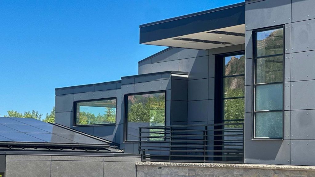 Modern house exterior with dark gray walls and large glass windows reflecting clear blue sky and greenery. Solar panels on roof. Sleek, minimalist design.