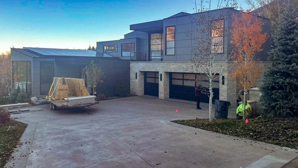 Modern two-story house with gray and stone exterior, large windows, and a spacious driveway. A trailer with construction materials is parked in front. Autumn trees add color.