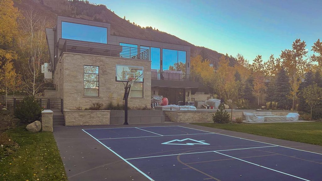 Modern house with large glass windows overlooks a backyard basketball court at sunset. Autumn trees surround the property, creating a tranquil setting.