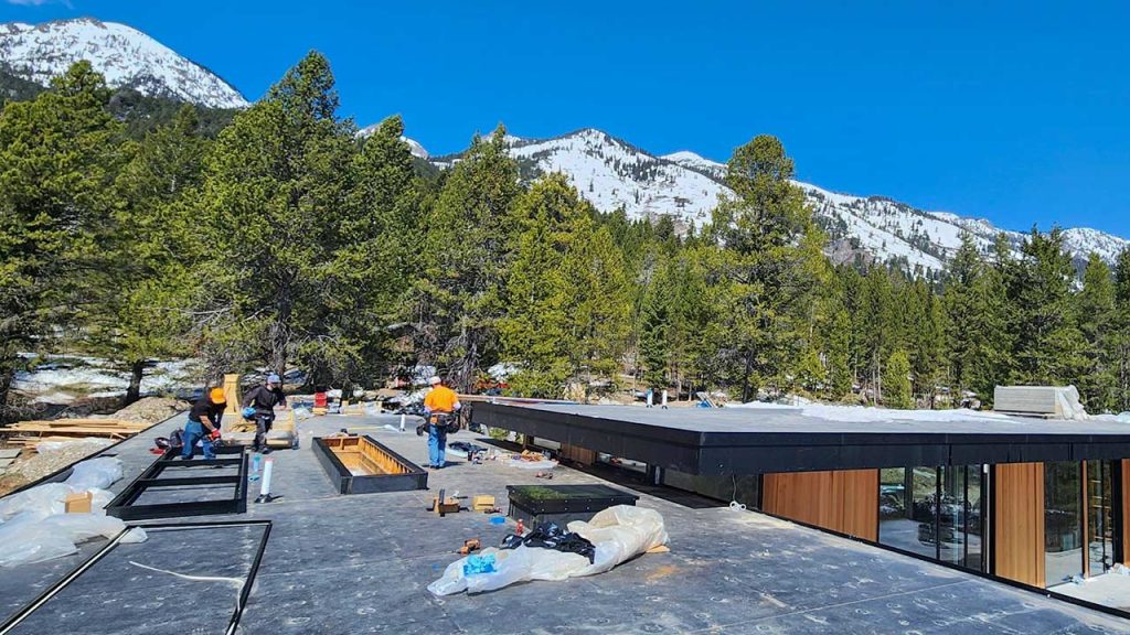 Two workers in orange safety gear work on a skylight frame assrmbly on a flat rooftop with snowy mountains and pine trees in the background. Clear blue sky enhances the serene setting.