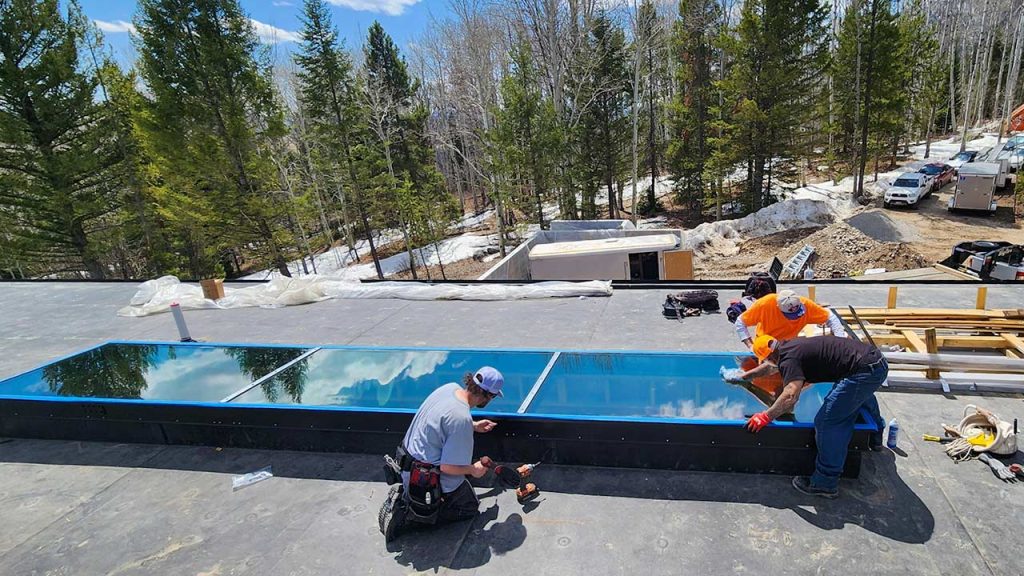 Three workers install a large glass skylight on a building surrounded by snow-dusted evergreen trees. The sky is clear, and sunlight reflects off the glass.