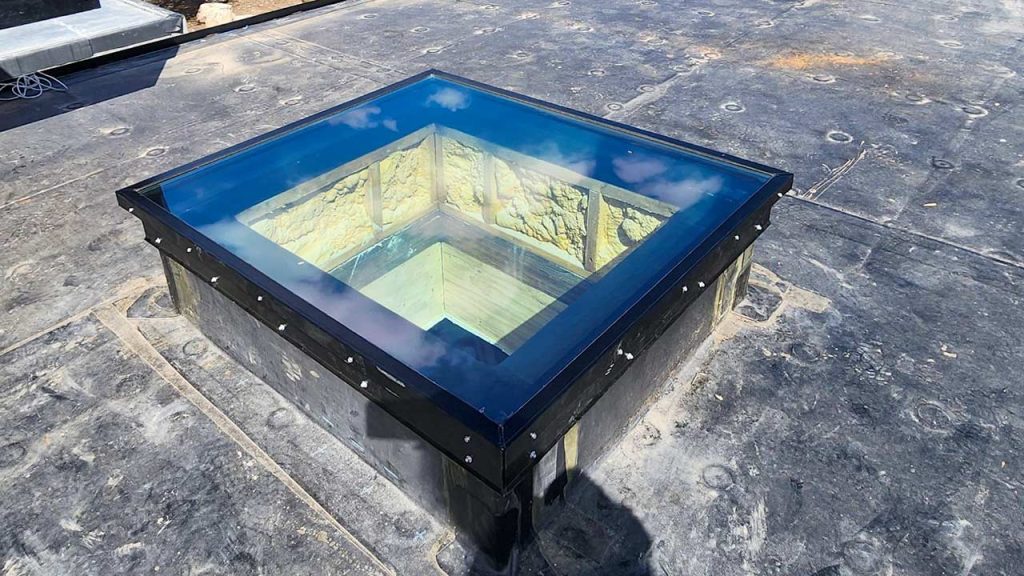 A square skylight with a reflective blue glass surface is set into a flat, gray rooftop. The skylight reveals a wooden shaft below, surrounded by stone walls.