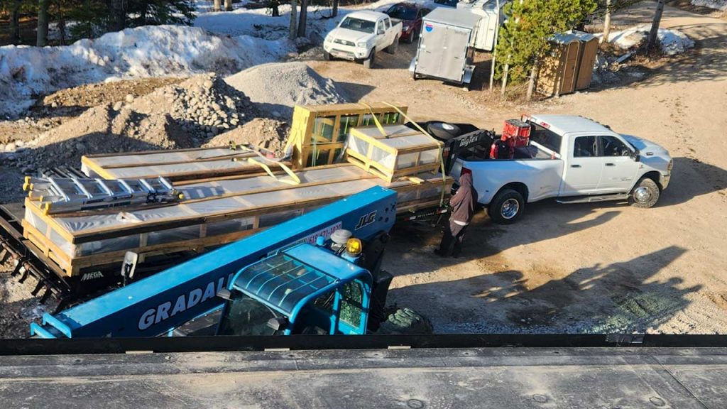 A construction site with a white pickup truck and a trailer loaded with skylight materials, including wooden frames and ladders. A blue forklift is ready to assist. Snow and trees are in the background.