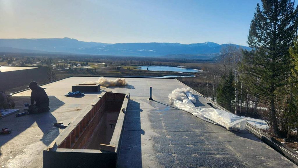 Rooftop view of open countryside with distant mountains, a lake, and tall trees. A person works on the roof in the foreground near a skylight curb, under clear skies.