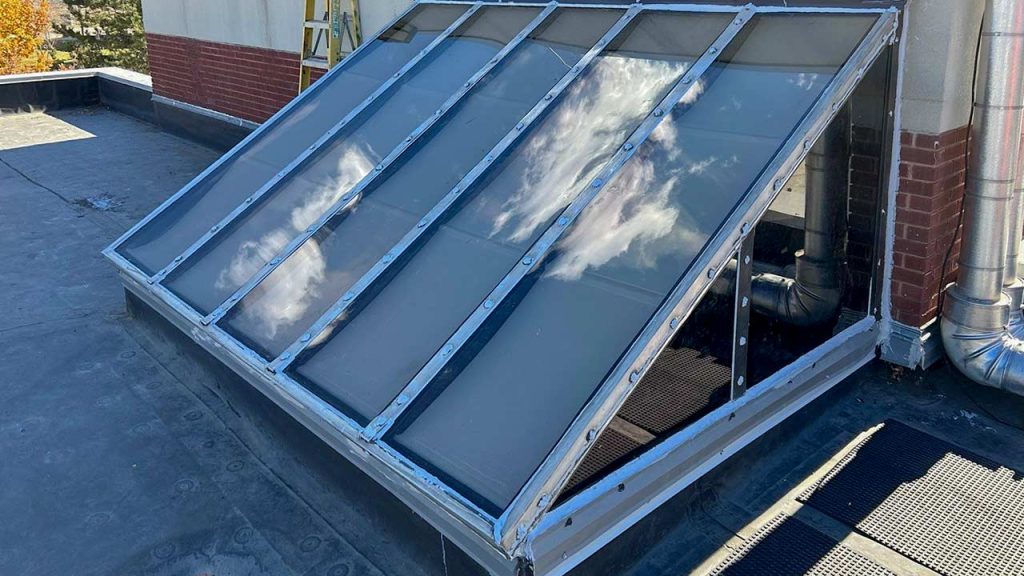 A rooftop skylight with a metal frame and polycarbonate panels reflects clouds against a blue sky. The setting is industrial with red brick and duct pipes nearby.