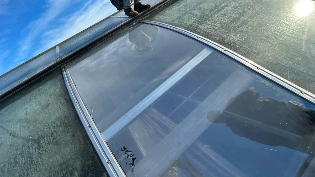 Person standing near a large, transparent, curved plastic skylight, with reflections of a blue sky. The scene conveys a sense of balance and focus.